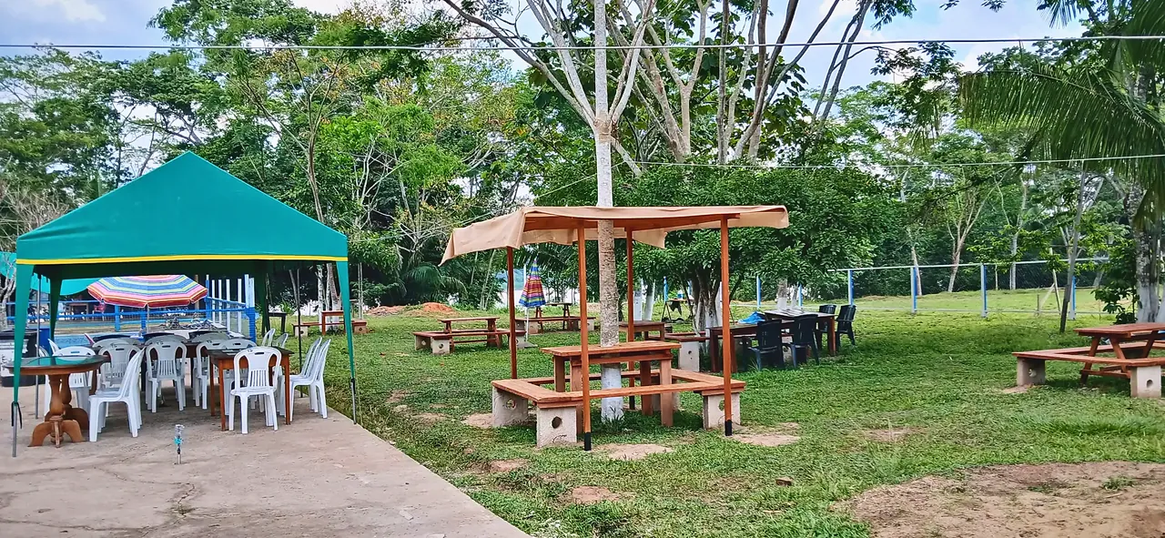 Jardín con una glorieta de madera con techo de lona y mesas de picnic bajo los árboles; a la izquierda, un toldo verde con mesas y sillas.