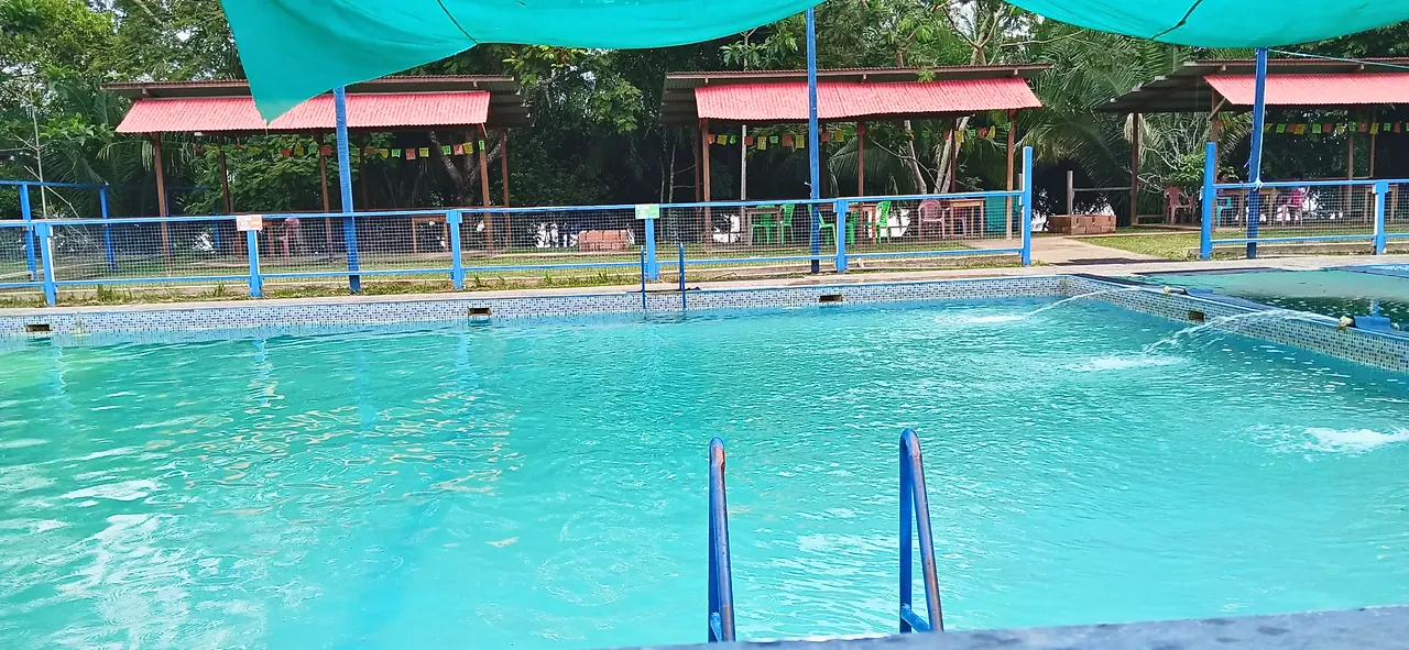 Piscina de agua turquesa con escalerilla azul y chorros de agua; al fondo, techos rojos con banderines entre los árboles.