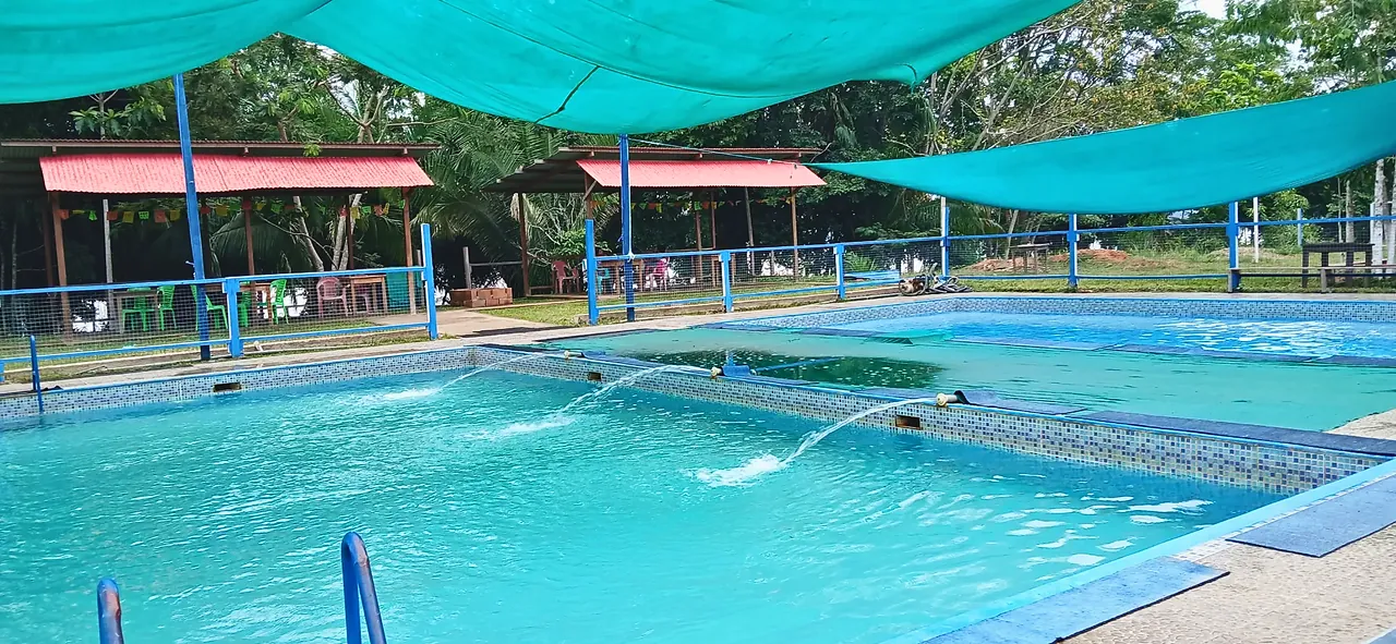 Piscina con tres chorros de agua bajo toldos de sombra; al fondo, zonas techadas con techos rojos.