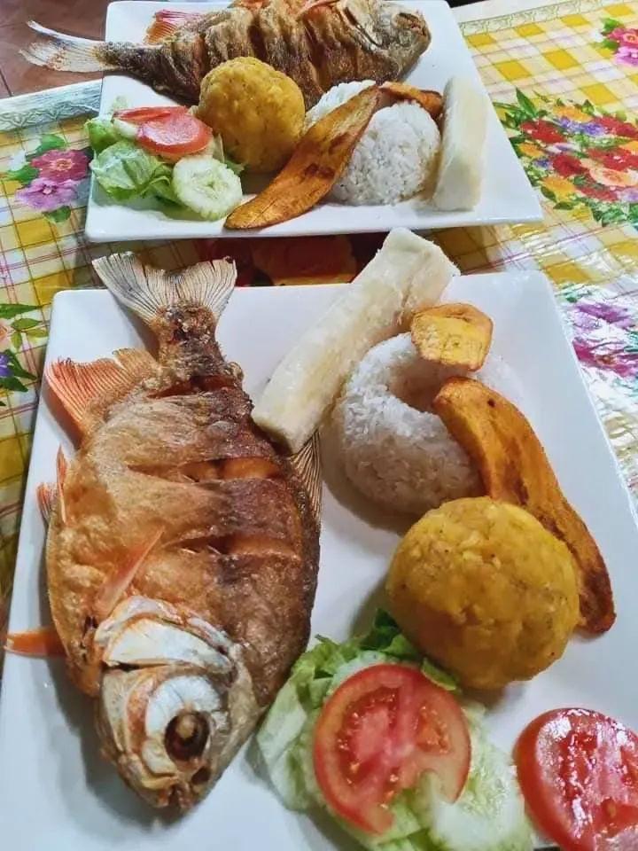 Dos platos de pescado frito entero con arroz, yuca, plátano frito, una bola dorada y ensalada de lechuga, tomate y pepino.