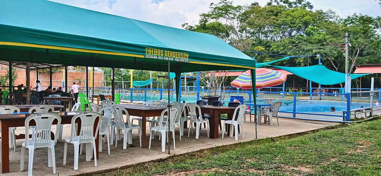 Toldo verde con mesas y sillas blancas junto a la piscina, donde se come a la sombra.